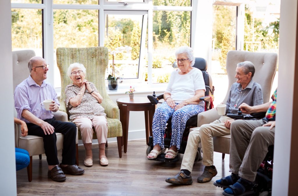 A group of seniors sitting in a semicircle in a common area, drinking tea or coffee and laughing