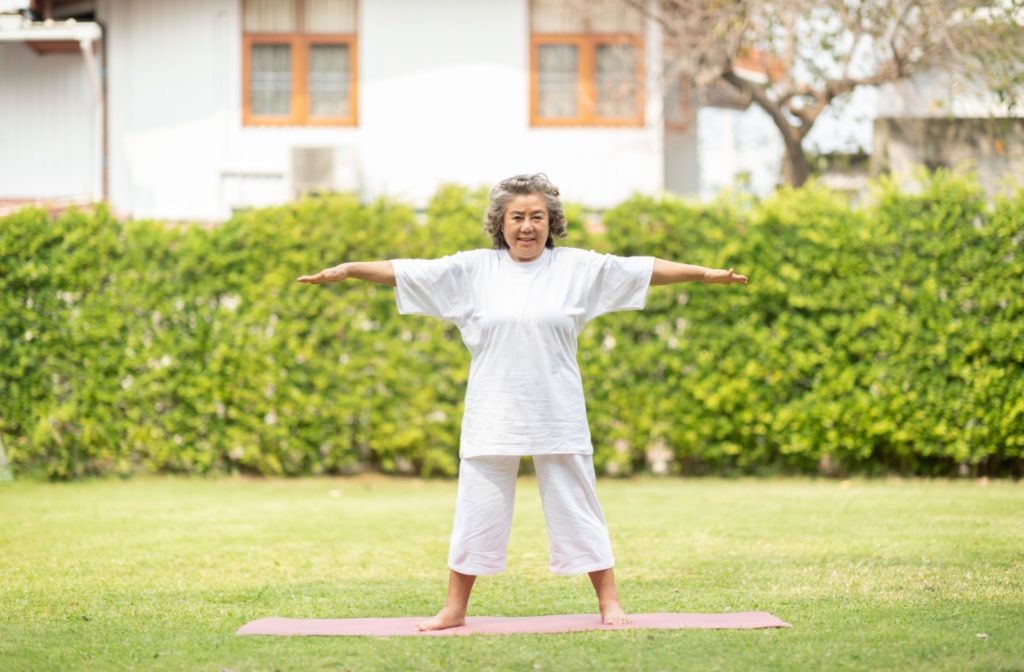 A senior stands—legs apart, arms outstretched—on a yoga mat outside.