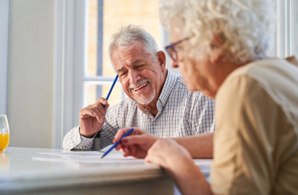 An older adult grins and tabs their forehead with a pen during a group puzzle activity in memory care